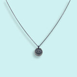 The Sterling Silver Evil Eye Medallion Necklace by Ornamental Things features a circular engraved eye pendant, shown against a light blue background.