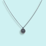 The Sterling Silver Evil Eye Medallion Necklace by Ornamental Things features a circular engraved eye pendant, shown against a light blue background.