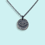 The Sterling Silver Evil Eye Medallion Necklace by Ornamental Things features an engraved eye with radiating lines on a round pendant, hanging from a blackened silver chain, displayed against a light blue background.