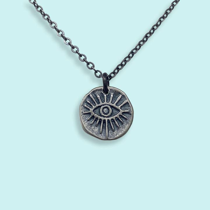 The Sterling Silver Evil Eye Medallion Necklace by Ornamental Things features a blackened silver chain and engraved round pendant, surrounded by rays, shown on a light blue background.