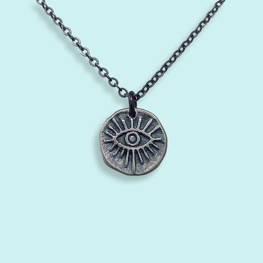The Sterling Silver Evil Eye Medallion Necklace by Ornamental Things features a blackened silver chain and engraved round pendant, surrounded by rays, shown on a light blue background.