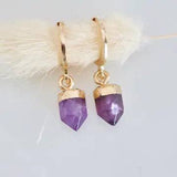 Mesa Blue’s Stone Bullet Amethyst Huggie Earrings feature 18K gold plated hoops with purple amethyst pendants capped in gold, shown against a light background.