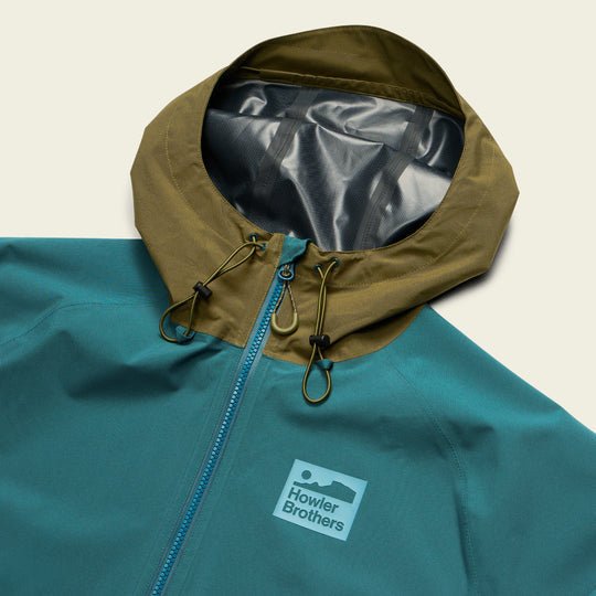 The Storm Splitter Rain Shell by Howler Brothers features a teal waterproof design, olive green hood, zip closure, and chest logo, shown against a plain background.
