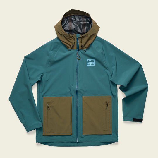 The Howler Brothers Storm Splitter Rain Shell features a teal and olive green waterproof design with two front zip pockets, a front zipper, and a light blue rectangular chest patch.