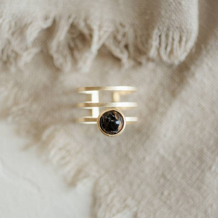 The Strata Ring Cooper Obsidian by CommonForm features three parallel gold bands and a round copper obsidian stone at the center, shown on textured beige fabric.