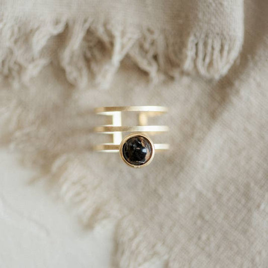The Strata Ring Cooper Obsidian by CommonForm features three parallel gold bands and a round copper obsidian stone at the center, shown on textured beige fabric.