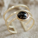 The Strata Ring Cooper Obsidian by CommonForm features three gold parallel bands and a central oval copper obsidian stone, beautifully bezel set and displayed on a neutral textured background.