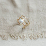 The Strata Ring Pearl by CommonForm, featuring three gold bands and a bezel-set pearl, is displayed on textured beige fabric with fringed edges.