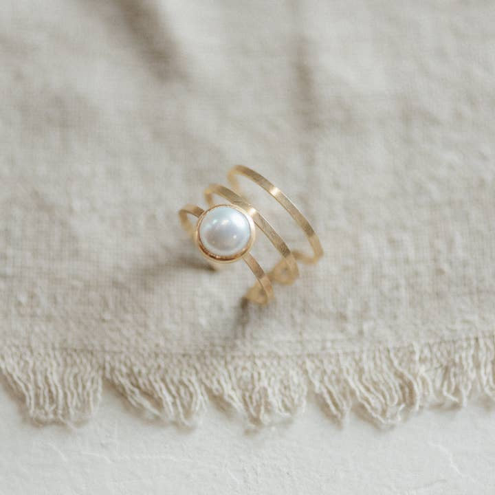 The Strata Ring Pearl by CommonForm, featuring three gold bands and a bezel-set pearl, is displayed on textured beige fabric with fringed edges.