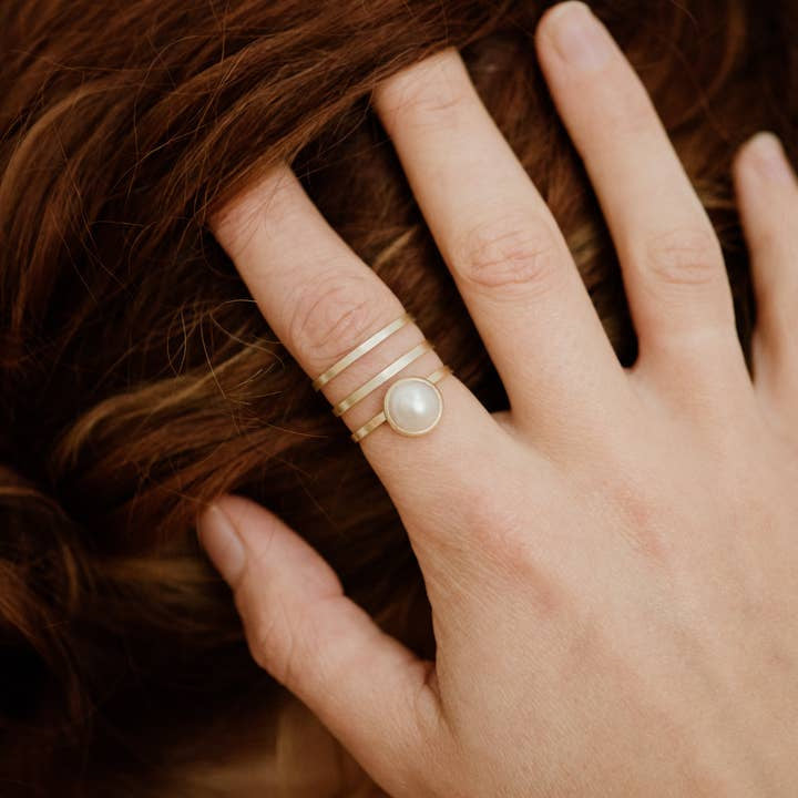 A hand adorned with four gold rings, including the Strata Ring Pearl by CommonForm—featuring a bezel-set pearl on an adjustable brass band—rests on wavy, light brown hair.