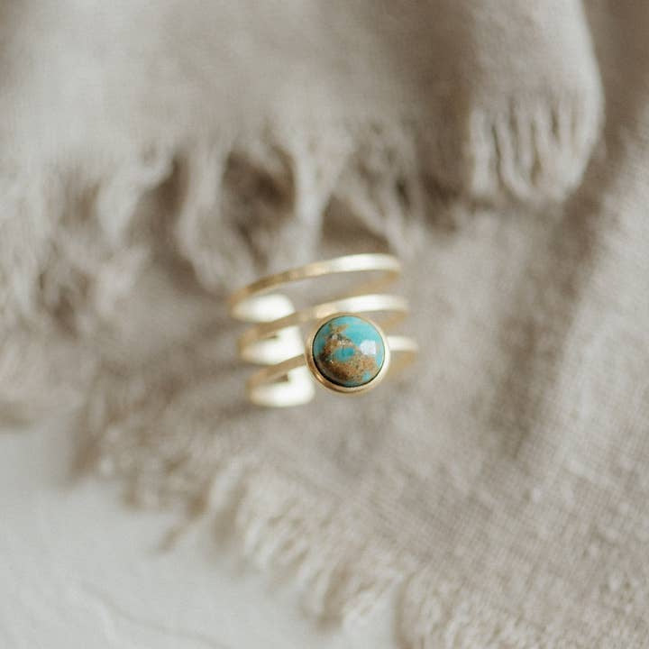 The Strata Ring Turquoise by CommonForm, featuring a bezel-set turquoise stone in a gold spiral design, rests on beige textured fabric.