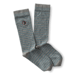 The Striped Logo Sock by Duck Head features gray and light blue stripes with a small round embroidered Duck Head logo patch near the top.