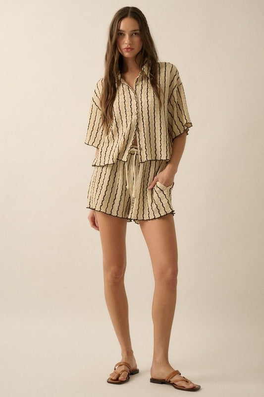 A woman stands against a neutral background wearing the Striped Textured Knit Button-Up by Promesa USA, a beige short-sleeve shirt with vertical stripes, coconut wood buttons, matching shorts, and brown sandals.