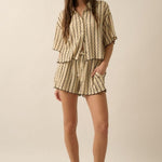 A woman stands against a neutral background wearing Promesa USA’s Striped Textured Knit Drawstring Shorts with a matching striped shirt and sandals, one hand in her shorts pocket.