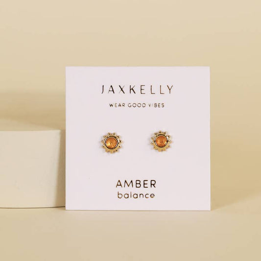 A pair of Sun Stud Earrings by JaxKelly featuring round amber hypoallergenic studs with gold sunburst settings is shown on a white card labeled "AMBER balance.