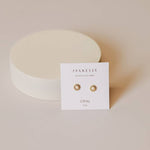 A pair of Sun Stud Earrings by JaxKelly, 18K gold plated studs with opal stones, shown on a branded "Opal Joy" card and placed before a round off-white object—elegant, tarnish-free jewelry from JaxKelly.