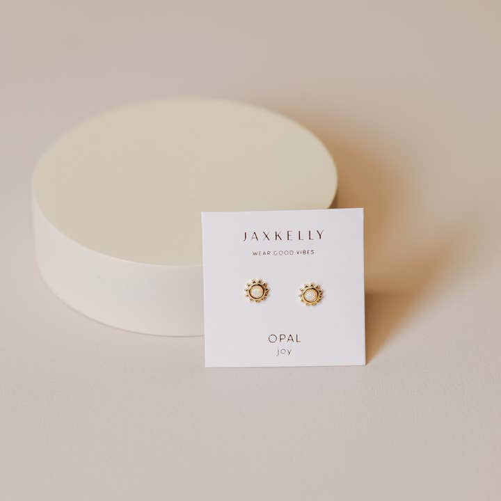 A pair of Sun Stud Earrings by JaxKelly, known for being hypoallergenic, rest on a white card labeled "Jax Kelly," displayed against a neutral background with a round white object behind them.