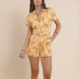 A woman stands against a beige background wearing the Sadie & Sage Sunset Beauty Zip Front Belted Romper and brown cowboy boots.