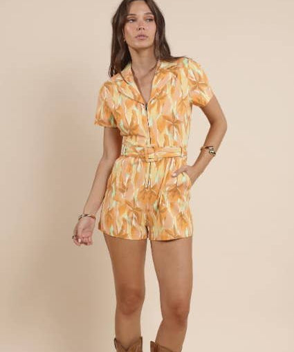 A woman wears the Sadie & Sage Sunset Beauty Zip Front Belted Romper and brown cowboy boots, standing against a plain beige background.