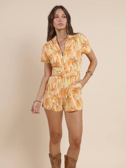 A woman stands against a beige background wearing the Sadie & Sage Sunset Beauty Zip Front Belted Romper and brown cowboy boots.