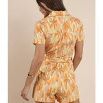 A woman faces away, modeling the Sadie & Sage Sunset Beauty Zip Front Belted Romper in an orange and yellow leaf print, crafted from a rayon-nylon blend, against a beige background.