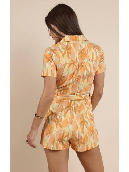 A woman faces away, modeling the Sadie & Sage Sunset Beauty Zip Front Belted Romper in an orange and yellow leaf print, crafted from a rayon-nylon blend, against a beige background.