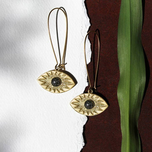 The Sunshine Eye Leverback Earrings by cameoko feature gold plating and black gemstone cabochon centers, displayed on a textured surface next to a green leaf.