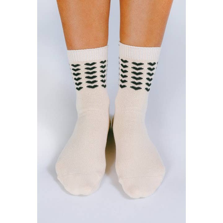 A pair of feet wear off-white Sweet Heart Ankle Socks by Tailored Union, featuring a black arrow pattern on the cuffs and made from premium combed cotton, shown against a plain background.