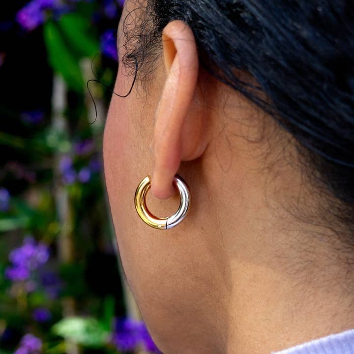 Close-up of a person wearing the Switch It Up Hoops by Peter and June—two-tone 18k gold plated and stainless steel earrings—with dark hair pulled back and blurred purple flowers in the background.