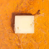 A bar of Tallow Soap by Health y Sol, made with all natural ingredients, rests in shallow orange-tinted water surrounded by bubbles and gentle ripples.