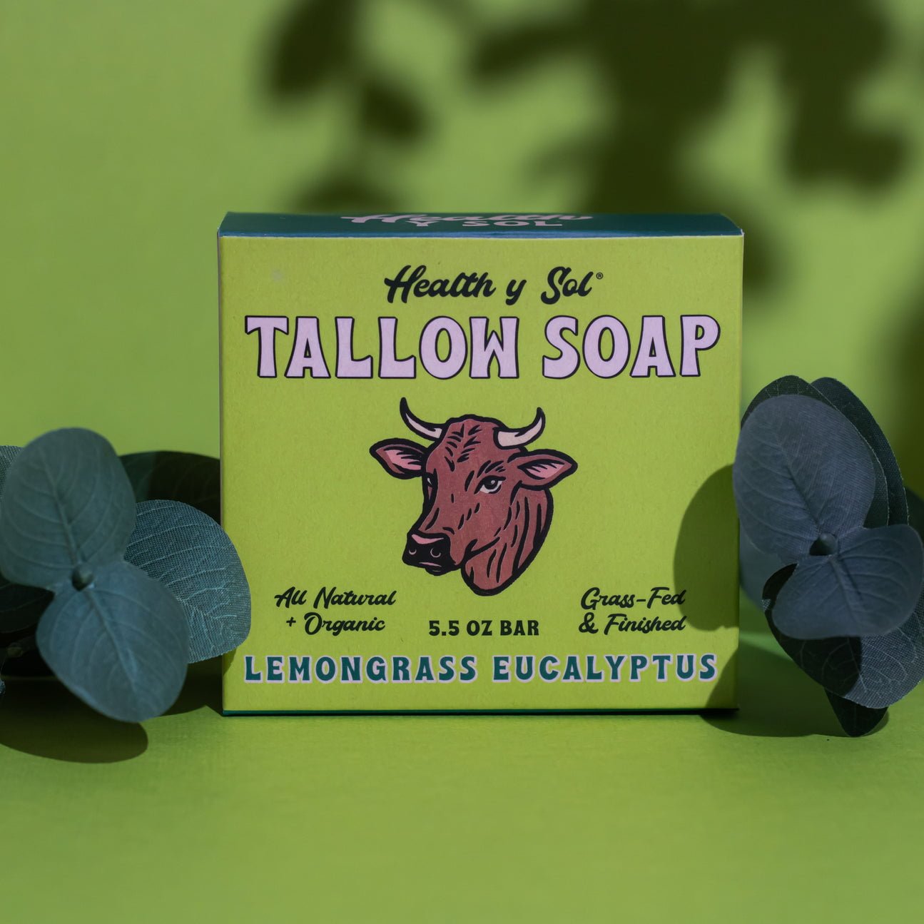 A box of Health y Sol Tallow Soap, made with all-natural ingredients and a lemongrass eucalyptus scent, sits on green beside blue flowers. The packaging shows a cow illustration and is gentle for eczema-prone skin. Brand: Health y Sol.
