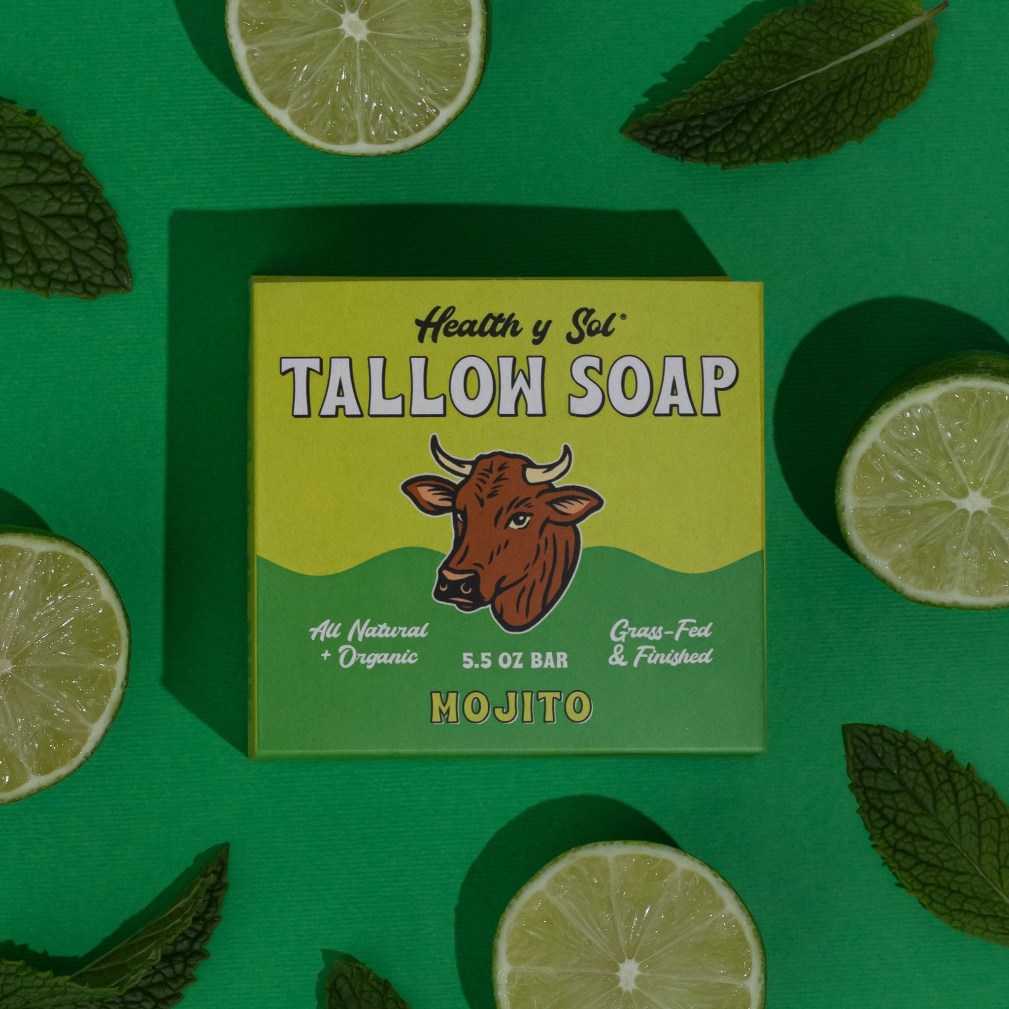 A box of Health y Sol Tallow Soap, Mojito scent, rests on a green surface with lime and mint. Made with natural ingredients, this organic soap may reduce acne and soothe eczema. The box displays a cow graphic. Brand: Health y Sol.