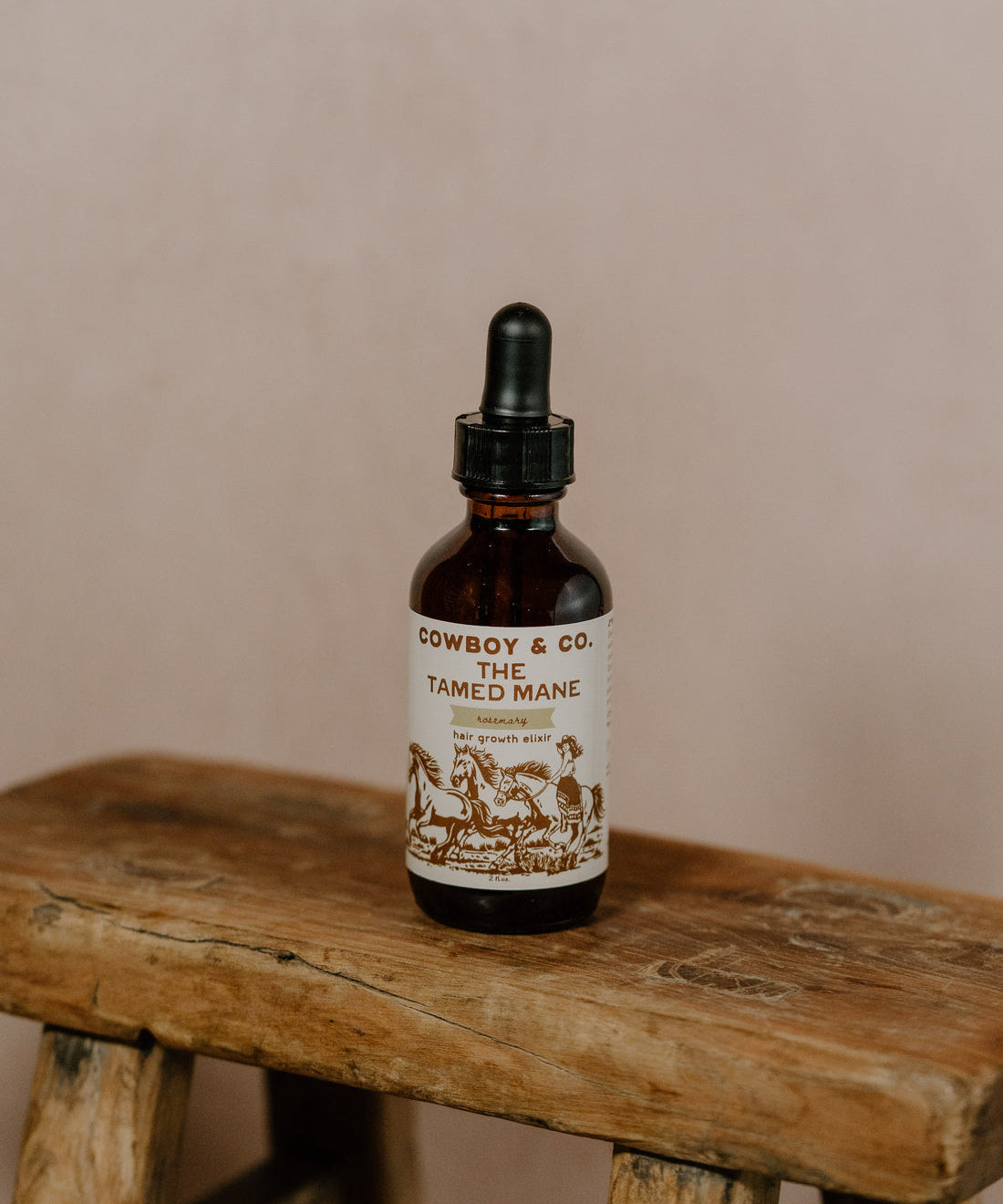A brown glass dropper bottle labeled "Cowboy & Co. TAMED MANE | Rosemary Hair & Scalp Elixir" rests on a rustic wooden stool, emphasizing its lightweight, natural hair care benefits against a neutral backdrop.