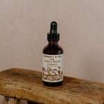 A brown glass dropper bottle labeled "Cowboy & Co. TAMED MANE | Rosemary Hair & Scalp Elixir" rests on a rustic wooden stool, emphasizing its lightweight, natural hair care benefits against a neutral backdrop.