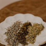 TAMED MANE | Rosemary Hair & Scalp Elixir by Cowboy & Co. sits on a white ceramic plate with dried herbs on wood—a perfect inspiration for natural hair and scalp care.