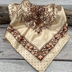 The Tan & Brown Paisley Wild Rag by Rodeo Drive, featuring brown paisley and floral patterns, is displayed on a wooden surface.