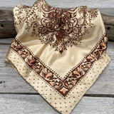 The Tan & Brown Paisley Wild Rag by Rodeo Drive, featuring brown paisley and floral patterns, is displayed on a wooden surface.