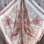 The Tan & Brown Paisley Wild Rag by Rodeo Drive features a cream base with brown and rust floral motifs plus four stylized peacocks, shown flat on wood. Perfect as a western scarf to add classic style to any outfit.