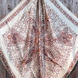 The Tan & Brown Paisley Wild Rag by Rodeo Drive features a cream base with brown and rust floral motifs plus four stylized peacocks, shown flat on wood. Perfect as a western scarf to add classic style to any outfit.