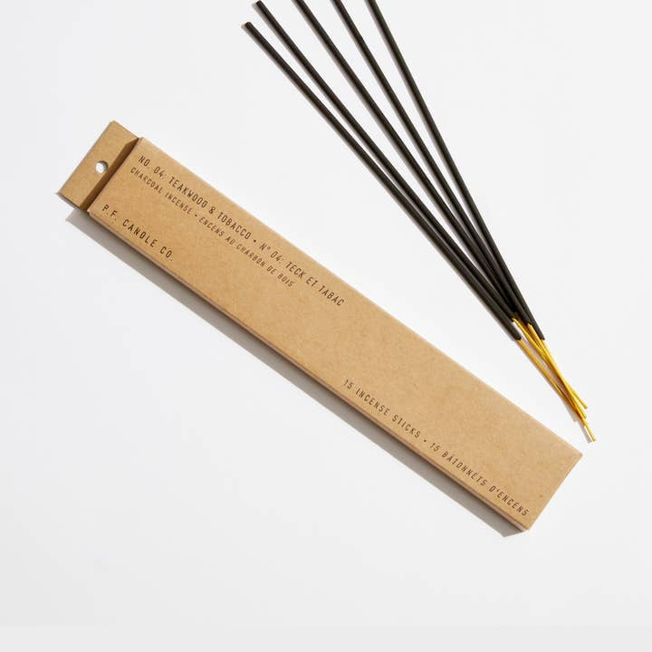A rectangular brown box labeled "P.F. Candle" with "Teakwood & Tobacco Incense Sticks," featuring the classic boyfriend scent, sits beside several yellow-tipped black incense sticks on a white surface.