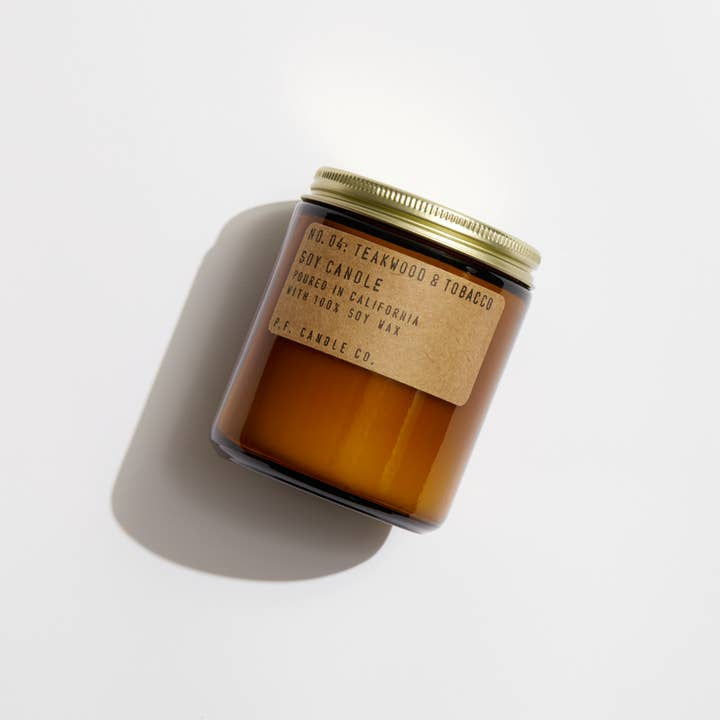 A brown amber jar candle with a gold lid and brown label reading “N0.4 Teakwood & Tobacco | P.F Candle” by P.F Candle, hand-poured and shown on a white surface.