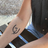 A person's forearm rests on a wooden table, featuring a Jenni Earle Temporary Tattoo with a minimalist Western horseshoe design and a small flower inside.