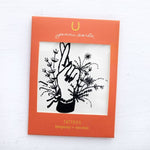 The Jenni Earle Temporary Tattoos feature a black line drawing of a hand with crossed fingers among flowers, framed in orange packaging labeled with the Jenni Earle brand. Non-toxic and easy to apply.