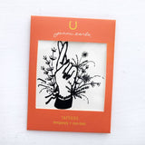 The Jenni Earle Temporary Tattoos feature a black line drawing of a hand with crossed fingers among flowers, framed in orange packaging labeled with the Jenni Earle brand. Non-toxic and easy to apply.