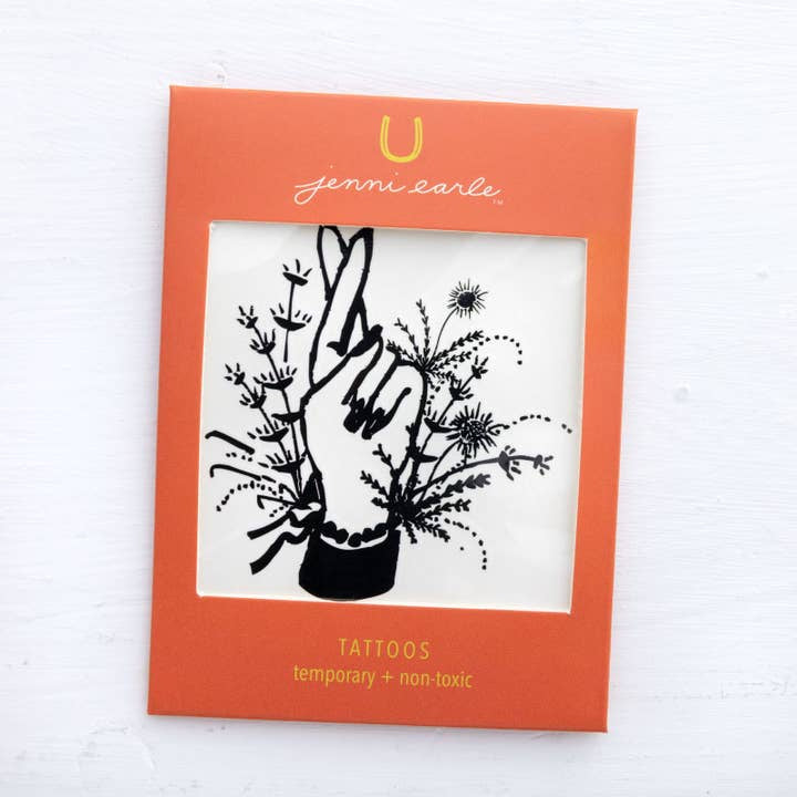 The Jenni Earle Temporary Tattoos feature a black line drawing of a hand with crossed fingers among flowers, framed in orange packaging labeled with the Jenni Earle brand. Non-toxic and easy to apply.