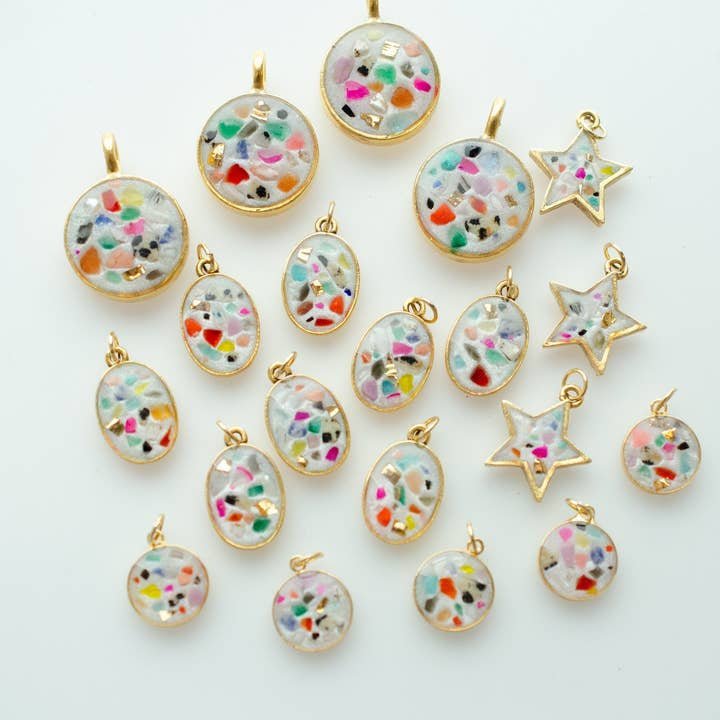 The Terrazzo Stone Necklace by Jill Makes features gold-framed resin pendants in oval, round, and star shapes with multicolored confetti accents and pearl inlay, displayed on a white surface.