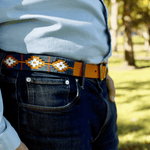 A person stands outdoors on grass, wearing jeans, a light blue shirt, and a Texas Monthly 50th Anniversary belt by Zilker Belts, made of Argentine leather. Trees are visible in the background.