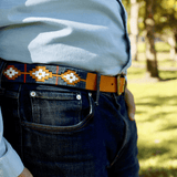 A person stands outdoors on grass, wearing jeans, a light blue shirt, and a Texas Monthly 50th Anniversary belt by Zilker Belts, made of Argentine leather. Trees are visible in the background.