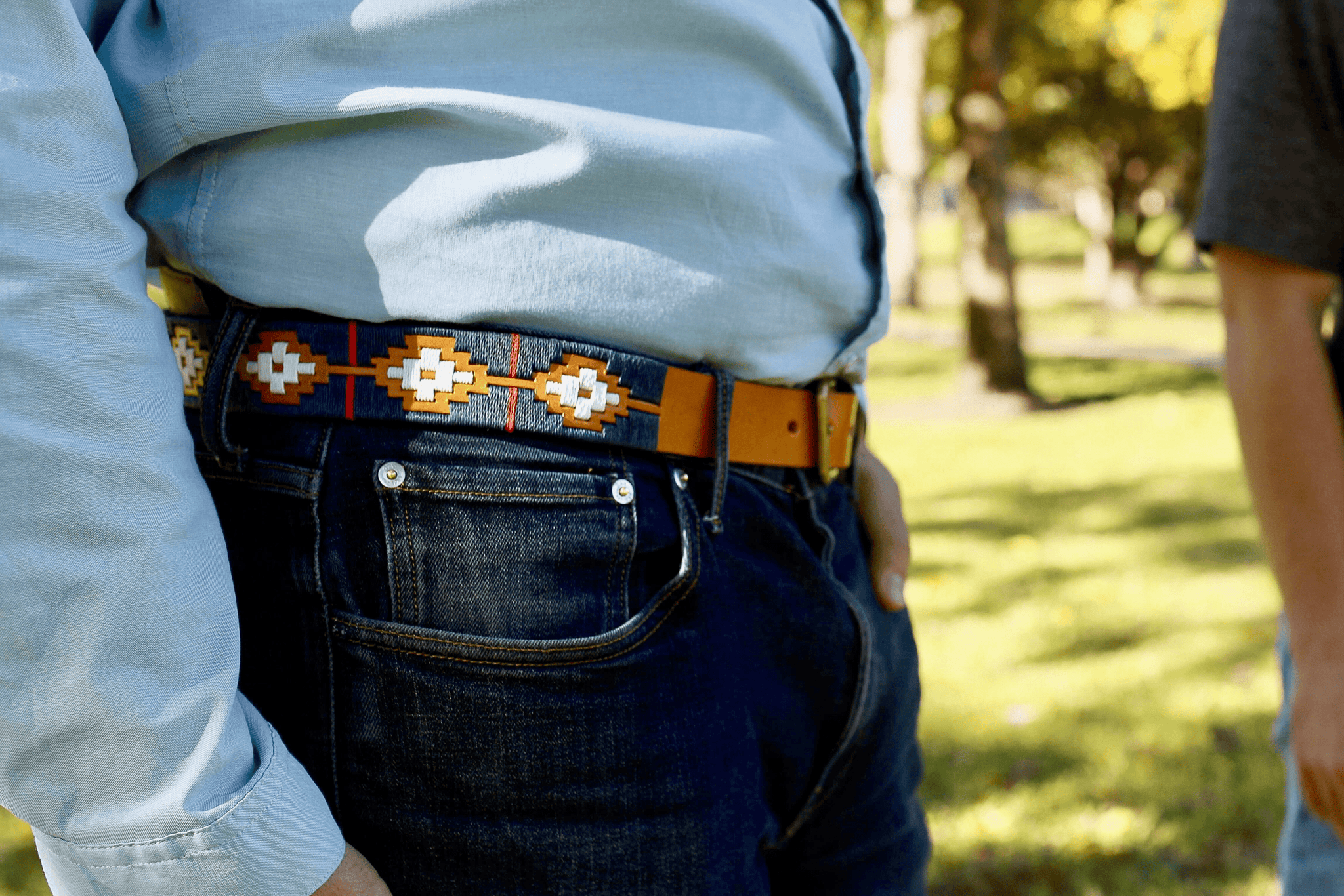A person stands outdoors on grass, wearing jeans, a light blue shirt, and a Texas Monthly 50th Anniversary belt by Zilker Belts, made of Argentine leather. Trees are visible in the background.
