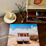 The "Texas Textures, Volume Two Book" by Kat Swansey rests on a table beside a teacup, decorative boot, and music CDs, its cover displaying a Kat Swansey photo of an abandoned Texas trailer.